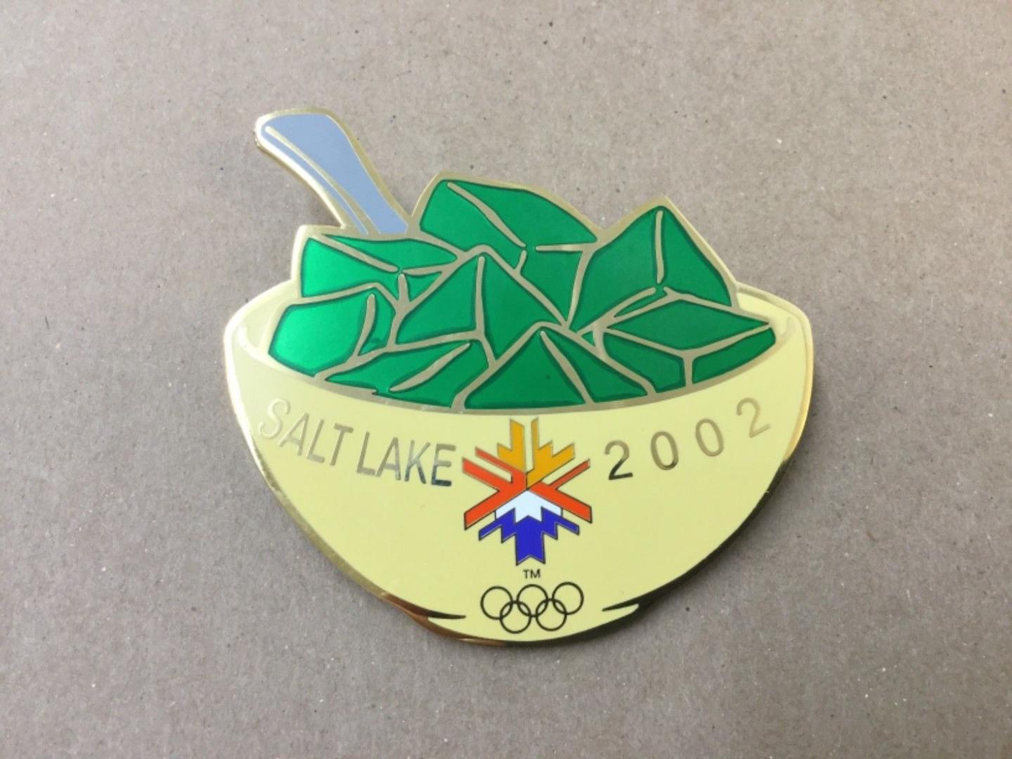 A commemorative pin from the 2002 Salt Lake City Winter Olympics shaped like a yellow bowl of green Jell-O with a spoon, featuring the Olympic rings and the official snowflake logo.