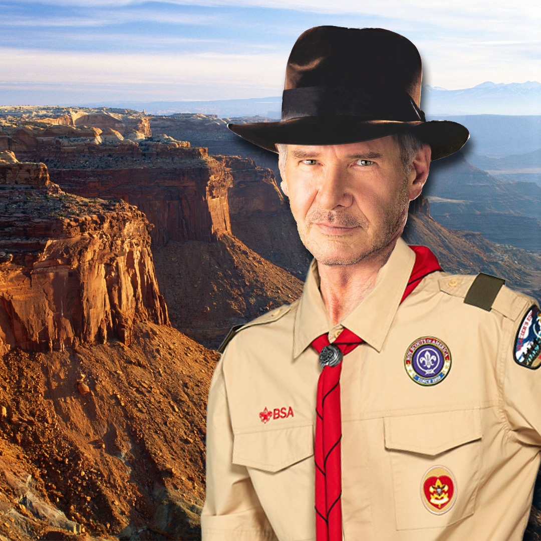 A digitally manipulated image of a man resembling Indiana Jones, wearing a tan Boy Scouts of America (BSA) uniform with various patches, a red neckerchief, and a black fedora. He has a confident expression and a rugged appearance. The background features a vast, rocky canyon landscape with reddish-brown cliffs under a bright blue sky.