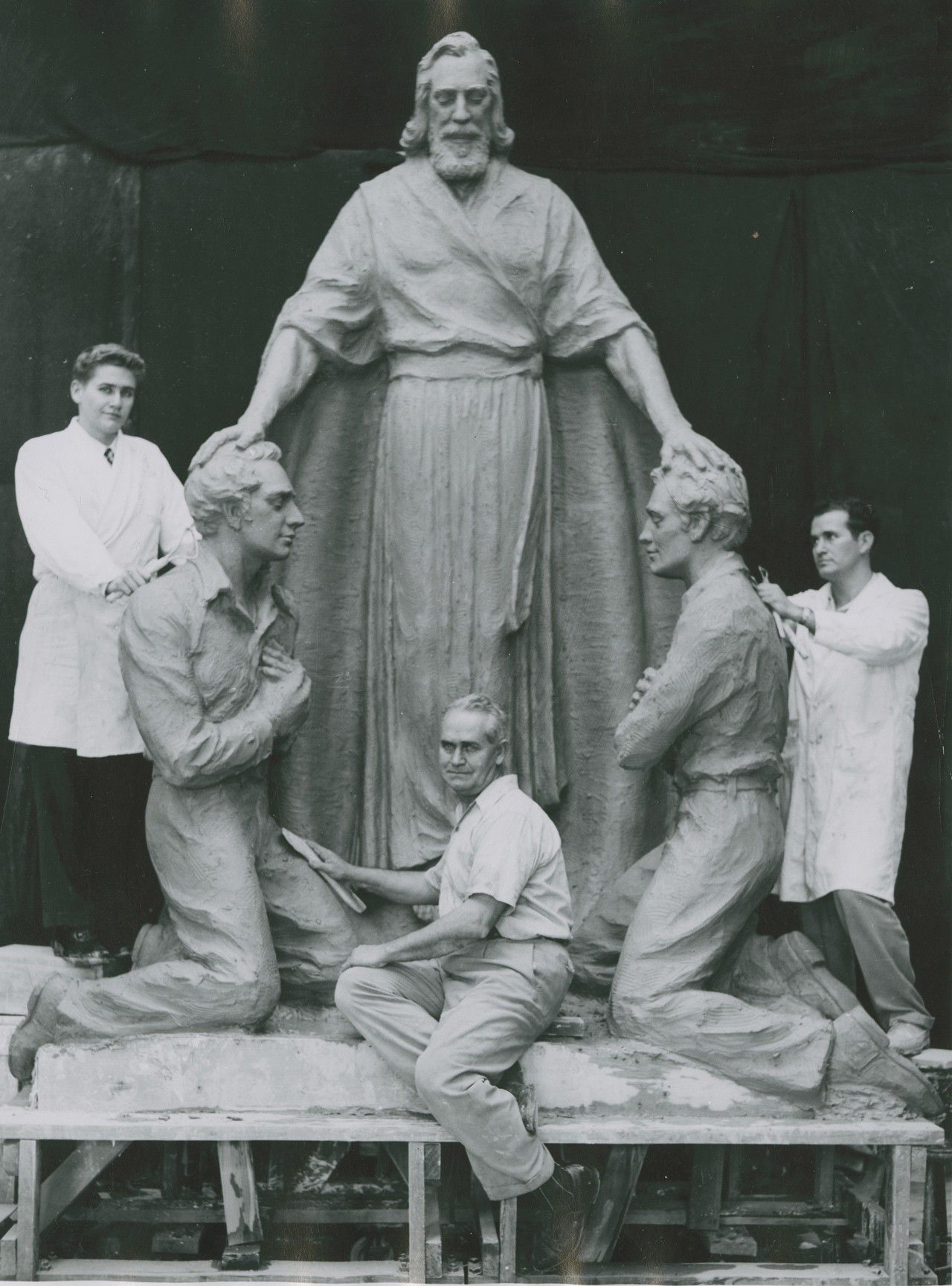 Three men pose with a large clay sculpture depicting a bearded figure standing with arms extended, placing his hands on the heads of two kneeling figures. The sculptor, Avard Fairbanks, sits in front of the statue holding sculpting tools, while two assistants in white coats stand behind the kneeling figures, working on the sculpture.