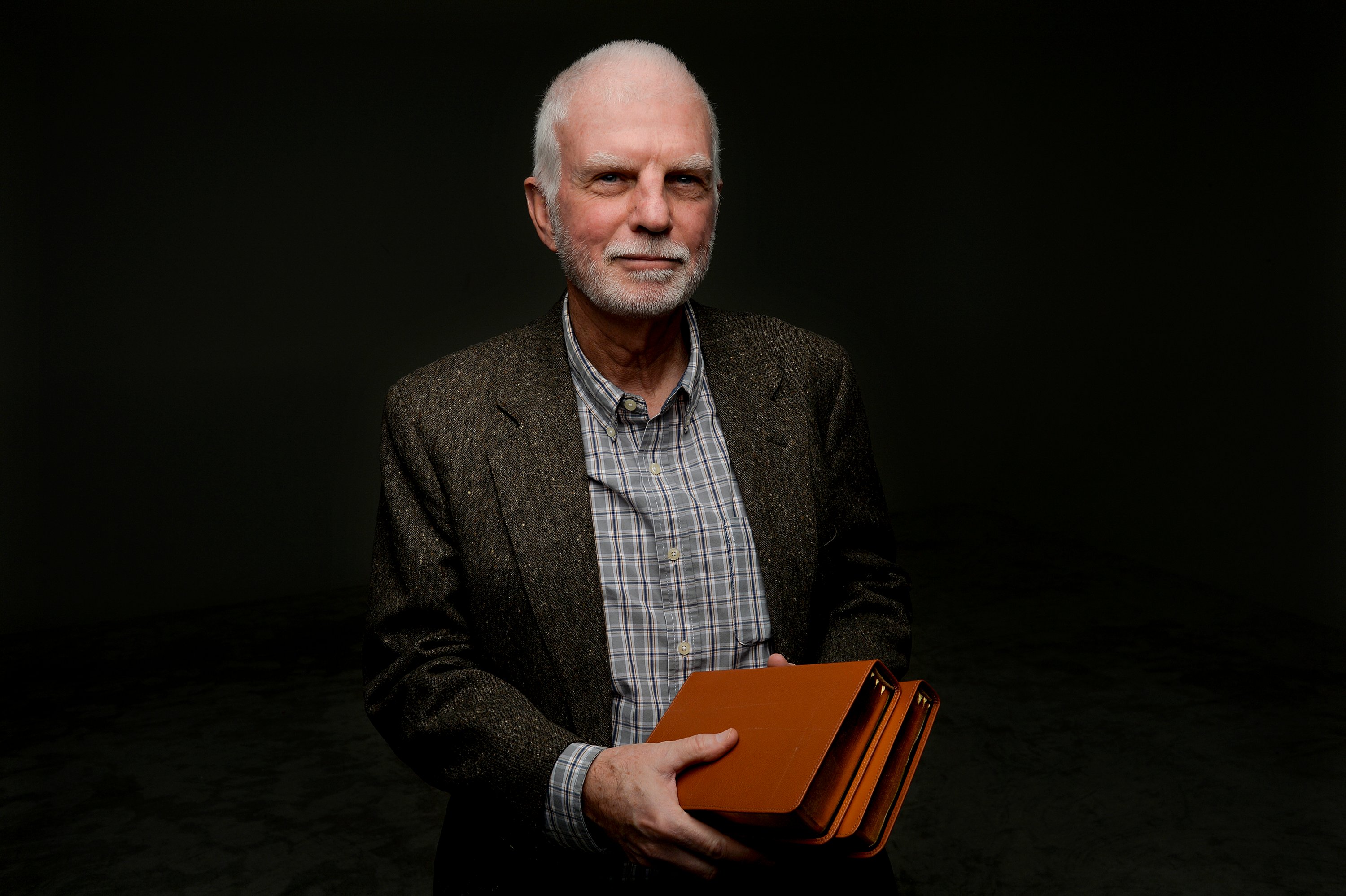 A photograph of a balding man with a gray beard, Denver Snuffer, holding two leather volumes of scripture against a black background.