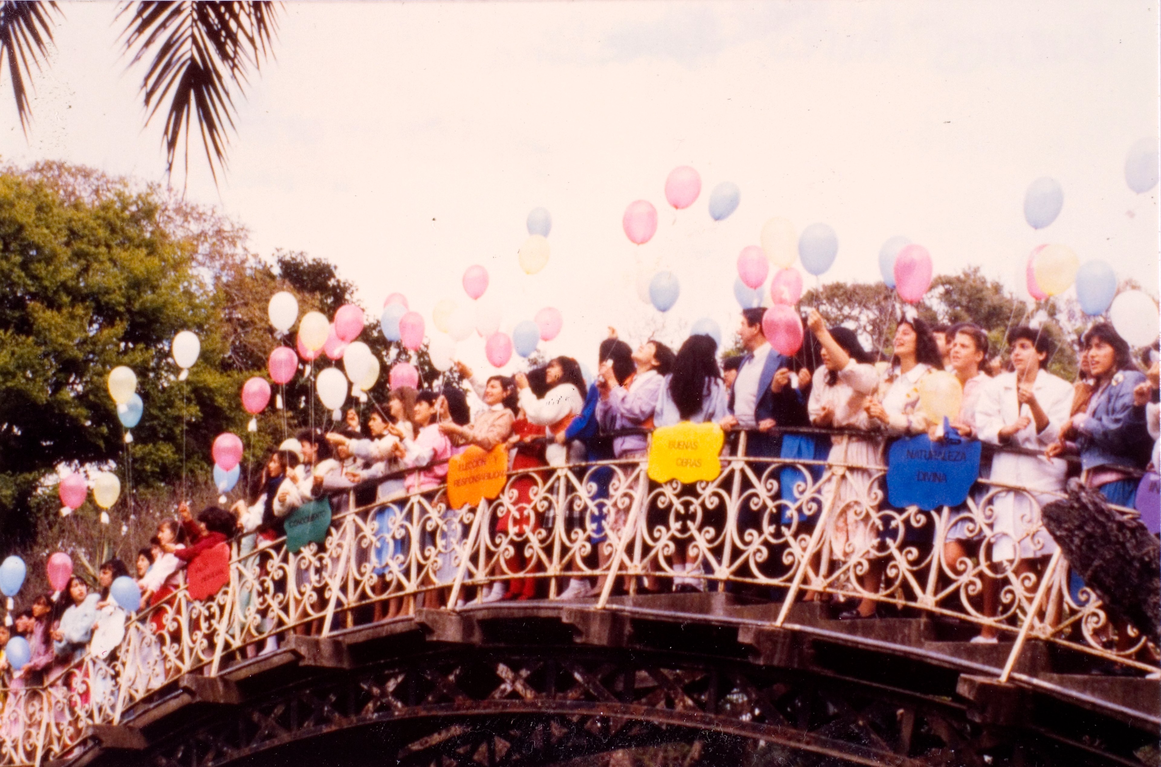 A large group of young women stand on an ornate metal bridge, releasing colorful helium-filled balloons into the sky. They are dressed in jackets and dresses, and some hold brightly colored signs with Spanish words such as "Buenas Obras" (Good Works) and "Naturaleza Divina" (Divine Nature). The background features trees and an overcast sky.