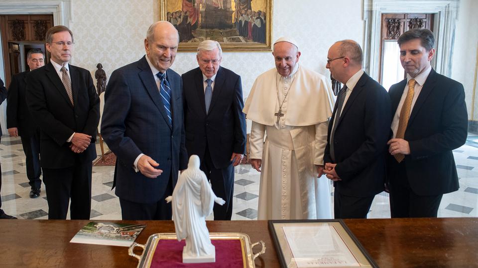 Pope Francis stands with the prophet and leaders of The Church of Jesus Christ of Latter-day Saints in a formal room with marble floors and ornate decor. They are gathered around a table, looking at a small white statue of Jesus Christ with outstretched arms, placed on a red cloth. Documents and a brochure are also on the table.