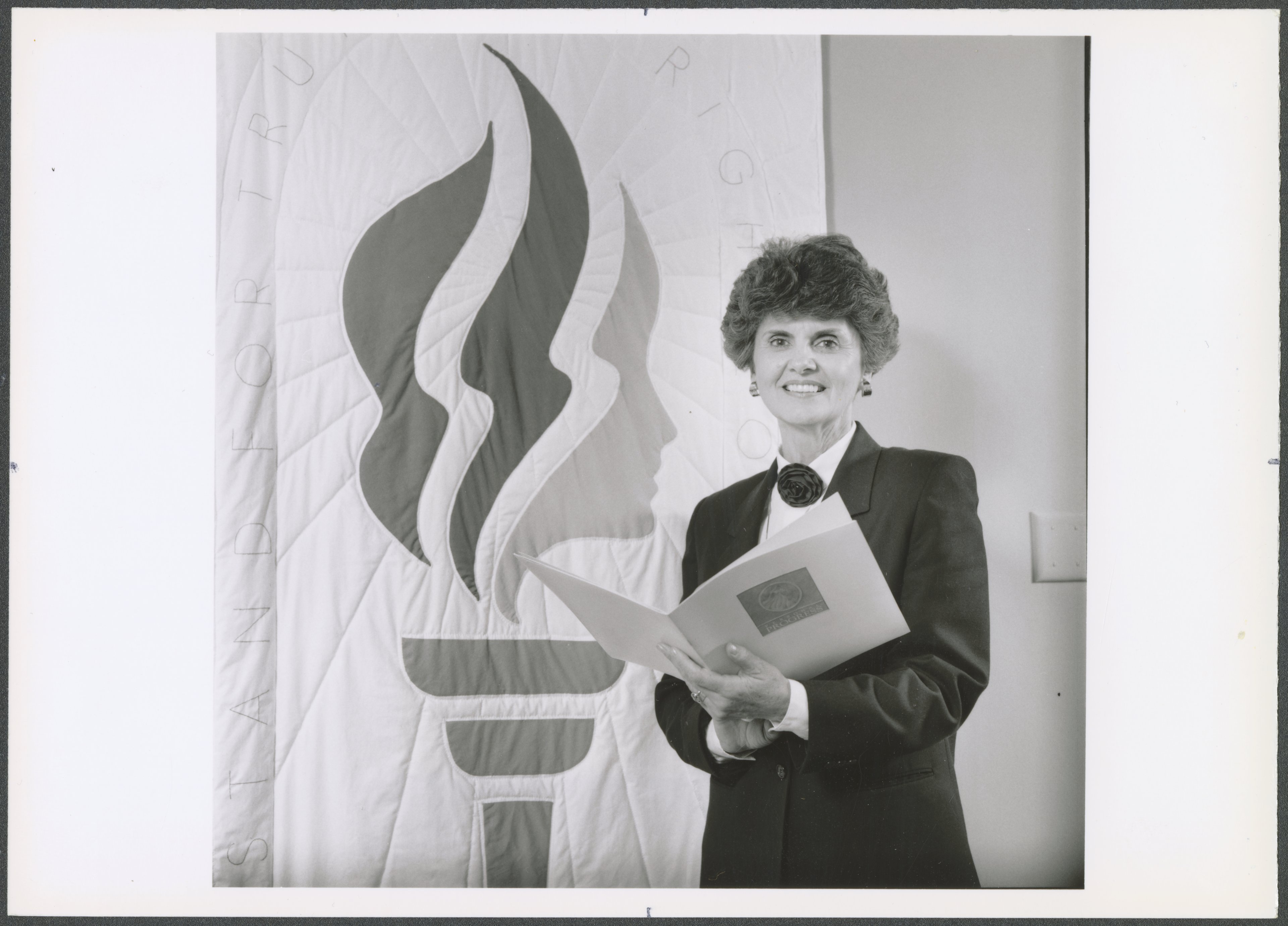 A black-and-white photograph of a woman with short, voluminous hair standing in front of a large quilted banner featuring a stylized torch and a profile silhouette. She is wearing a dark blazer with a white blouse and a decorative bow at the collar. She is holding an open booklet with an emblem on the cover. The background features a plain wall with a light switch visible.