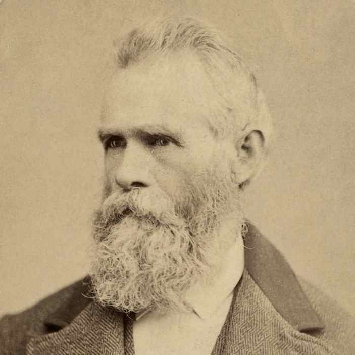 A sepia-toned vintage photograph of an older man with a full, bushy white beard and short, neatly combed hair. He has deep-set eyes gazing slightly to the side. He is wearing a dark, textured suit jacket over a white collared shirt. The background is plain and neutral.