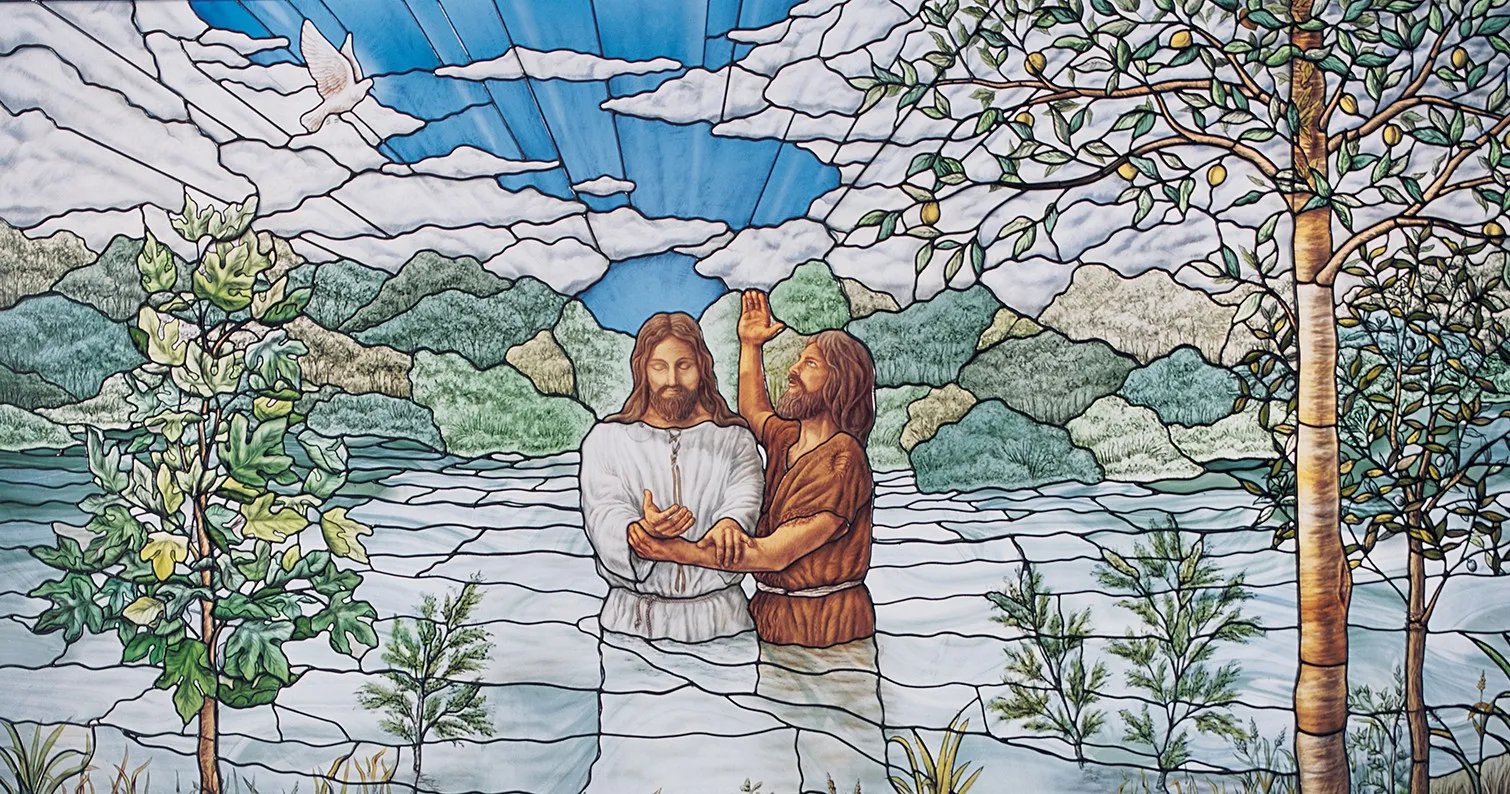 A stained glass artwork depicting John the Baptist baptizing Jesus in the Jordan River. Jesus, dressed in white, stands in the water with his hands gently clasped, while John, wearing a brown robe, raises his hand in blessing. The background features rolling green hills, a bright sky with rays of light streaming down, and a white dove symbolizing the Holy Spirit. Trees and plants frame the scene.