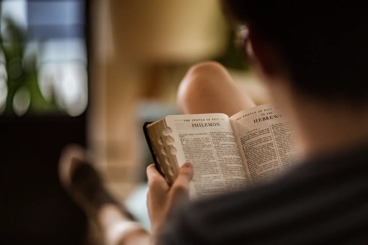 A person reading an open Bible, with the text visible from the books of Philemon and Hebrews. The reader is sitting in a relaxed posture with one leg propped up, and the background is softly blurred, showing an indoor setting with natural light filtering through a window.