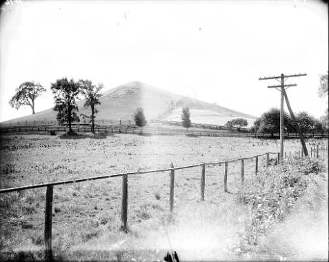 Uma fotografia em preto e branco de 1907 da Colina Cumora, em Nova York, mostrando uma encosta gramada com um caminho cercado por árvores, um pasto cercado em primeiro plano e postes de madeira na lateral.