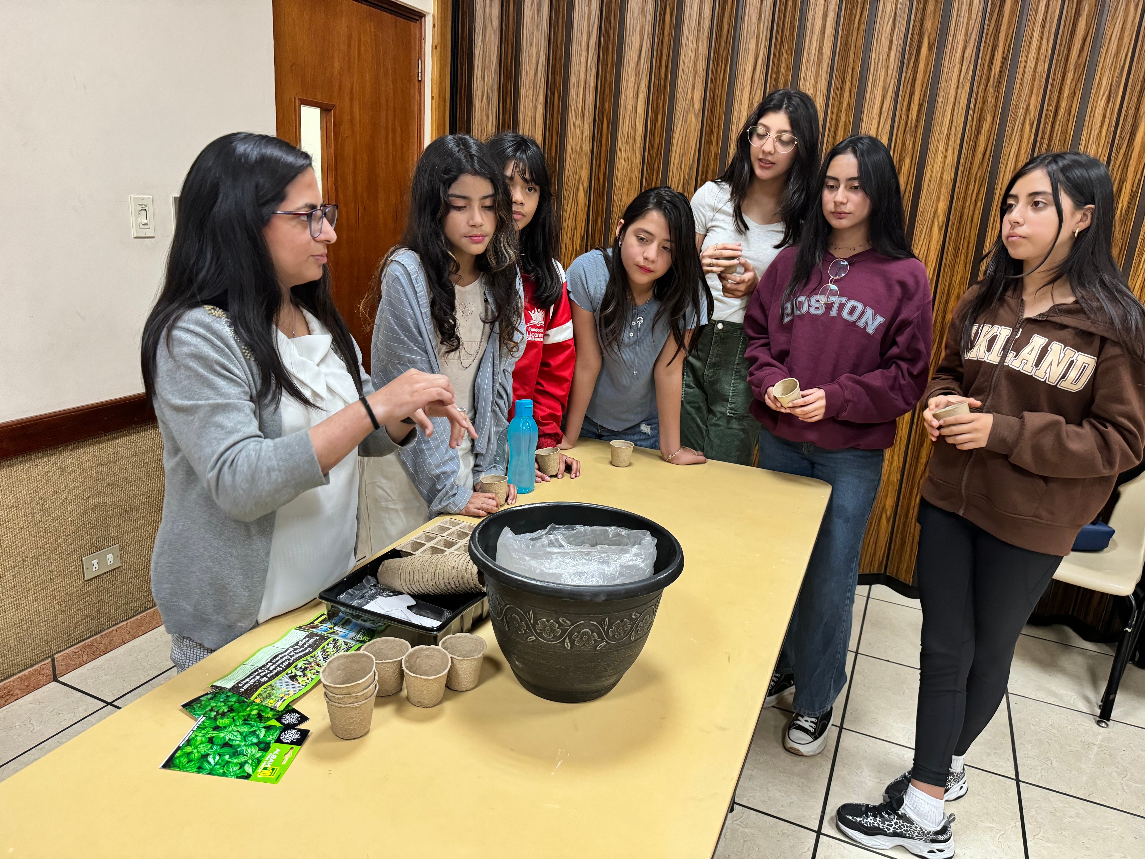 A group of young girls gathers around a table as a woman with long dark hair and glasses demonstrates a gardening activity. The table is covered with biodegradable pots, seed packets, a large decorative planter, and other gardening supplies. The girls, dressed in casual clothing including hoodies and jackets, watch attentively, some holding small pots. The setting appears to be an indoor classroom or community center with wooden paneling on the walls and a beige table.