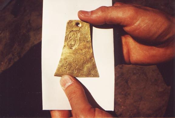 Een foto van twee handen die de laatst bekende Kinderhook-plaat vasthouden, een klein klokvormig stuk messing dat een vervalsing van een oud artefact was