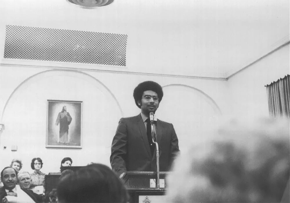Sur une photographie en noir et blanc, Darius Gray, conseiller d’origine dans la présidence du Groupe Genesis des saints des derniers jours, une organisation pour les membres afro-américains de l’Église, prend la parole lors de leur première réunion en 1971, avec une image de Jésus-Christ sur le mur derrière lui.